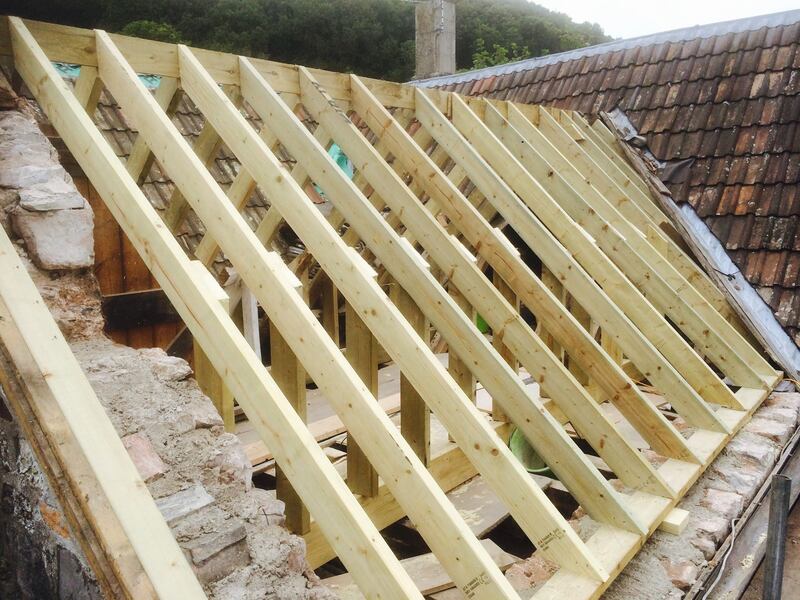New timber roof rafters being installed on top of an old stone wall, with existing red tile roofing visible behind.