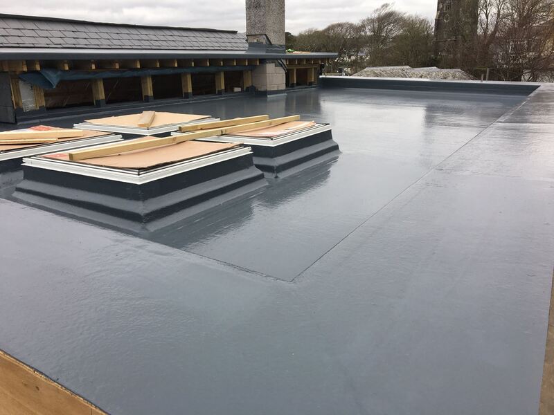 A large grey GRP fibreglass flat roof on a building with two raised timber roof-light upstands, seen against a church tower and trees in the background.
