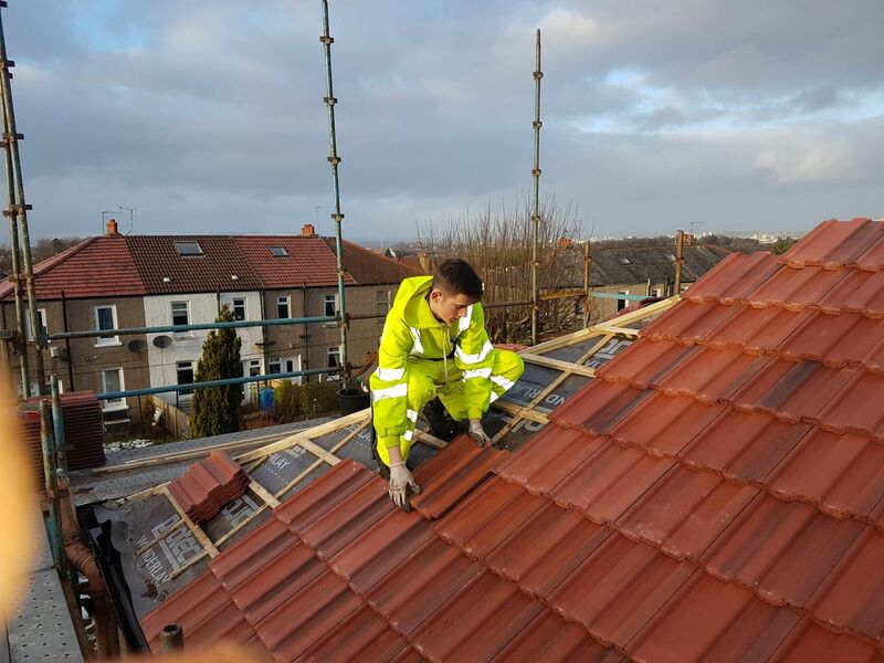 A roofer in high-vis clothing kneels on a partially tiled red pitched roof, fitting new clay tiles beside scaffolding overlooking terraced houses.